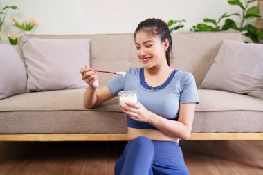 Yetişkin Asyalı kadın, oturma odasında sağlıklı yaşam için besleyici yoğurt atıştırmanın keyfini çıkarıyor. Kendi başının çaresine bakıyor..