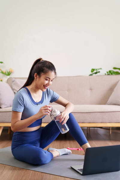 Взрослая азиатка в спортивном костюме отдыхает после домашней тренировки. Она улыбается, когда увлажняется из бутылки с водой возле ноутбука во время онлайн-упражнений. Здоровье и здоровье.
