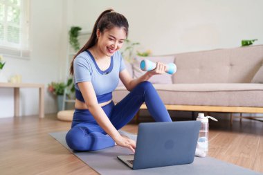 Gülümseyen Asyalı kadın, oturma odasındaki sağlıklı yaşam tarzı aktivitelerinde halterli halterlerle evde antrenman yapıyor.