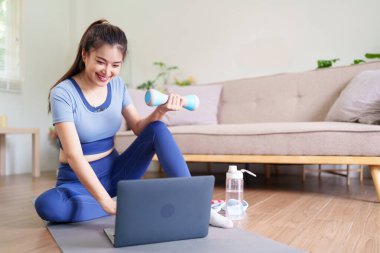 Yetişkin Asyalı kadın evde dizüstü bilgisayarlı halterle çalışmaktan hoşlanıyor. Sağlık hedeflerine ulaşmak için çevrimiçi fitness programını takip ediyor. Sağlığı motive eder.