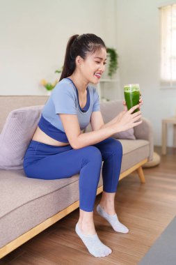 Aktif giysili gülümseyen Asyalı genç kadın kanepenin üzerinde besleyici yeşil smoothie 'nin keyfini çıkarıyor. Sağlıklı yaşam ve evde sağlıklı beslenmeyi destekliyor.