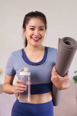 Gülümseyen Asyalı yetişkin kadın yoga minderi ve su şişesi tutarak antrenmana hazırlanıyor.