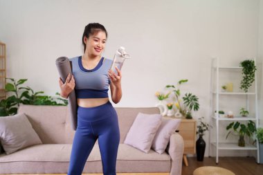 Gülümseyen Asyalı yetişkin kadın yoga antrenmanı için hazır. Paspas ve su şişesi sağlıklı sağlık ve sağlık yolculuğunu teşvik ediyor.