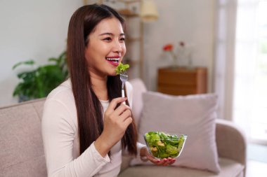 Neşeli Asyalı kadın taze yeşil salatanın koltukta rahatça oturmasından hoşlanıyor. Sağlıklı beslenme evde refahı ve dengeli bir yaşam tarzını teşvik eder..