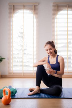 Yetişkin Asyalı kadın yoga minderinde akıllı telefon kullanarak gülümsüyor. Spor aletleri yakında, evde sağlıklı bir yaşam tarzını sembolize ediyor..