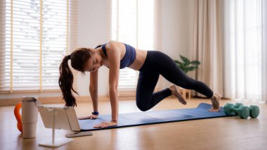 Asyalı yetişkin kadın güçlü bir ev egzersizi yapıyor. Sanal fitness koçluğu için bir tablet kullanıyor. Genel olarak sağlıklı olmayı teşvik ediyor..
