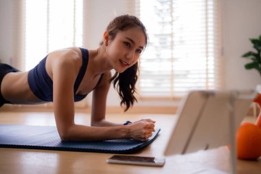 Asyalı kadın iç mekanda kalas egzersizi yapıyor. Çekirdek gücünü arttırıyor. Rehberlik için tablet kullanarak gülümsüyor..