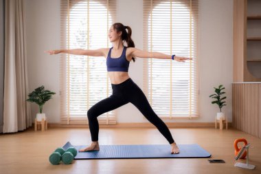 Sağlıklı yetişkin Asyalı kadın evinde savaşçı yoga pozu veriyor. Kişisel refah ve güç artışı için dikkatli bir fitness aktivitesine girişir..