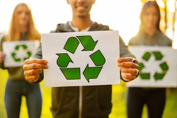 Midsection of young man holding placard with recycling symbol. Environmental volunteers in team standing at park. They are protecting nature.