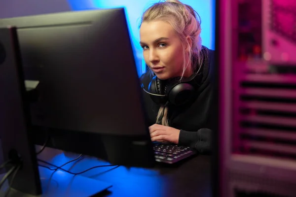 Professional blonde e-sport gamer woman with headphones playing online video game on her gaming workstation PC. Room with blue and pink colored LED lights.