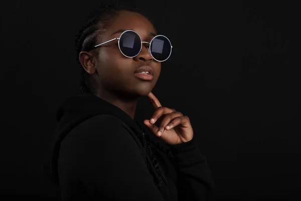 Dark portrait of confident and cool young african woman with dark skin wearing round sunglasses and sitting in a chair. High quality low-key studio portrait.