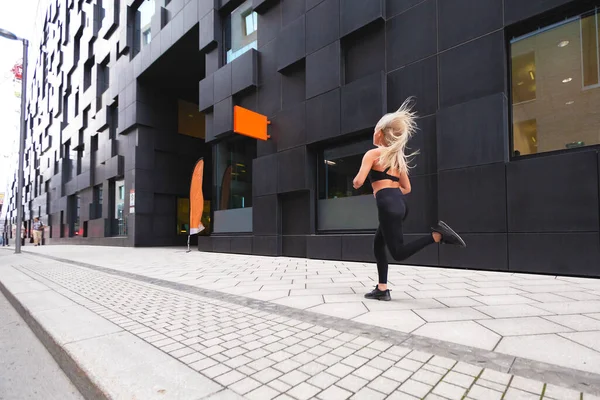 Siyah spor kıyafetli genç bir kadın minimalist şehir ortamında hızla koşuyor. Spor saati olan bir kadın..