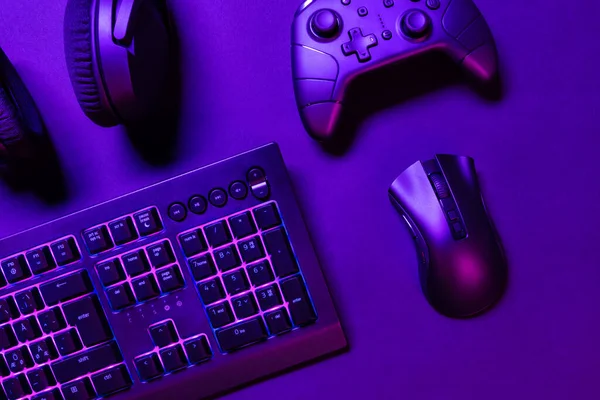 用鼠标和绿色发光键盘在明亮的紫色桌子上俯瞰游戏控制器和耳机 — 图库照片