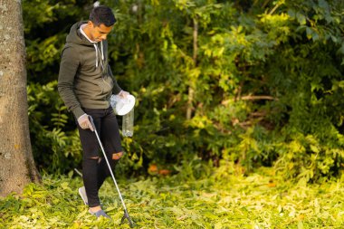 Güneşli bir günde parkta çimleri temizlemeye kendini adamış bir genç. Çevreyi kirlilikten koruyan sorumlu gönüllü. Elinde çöp ve kapkaççı tutuyor..