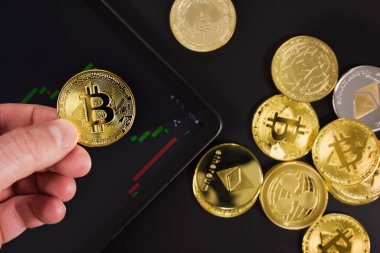 Siyah arka planda çeşitli kripto para birimleriyle altın Bitcoin BTC 'yi dijital tablet üzerinde tutan bir adam.