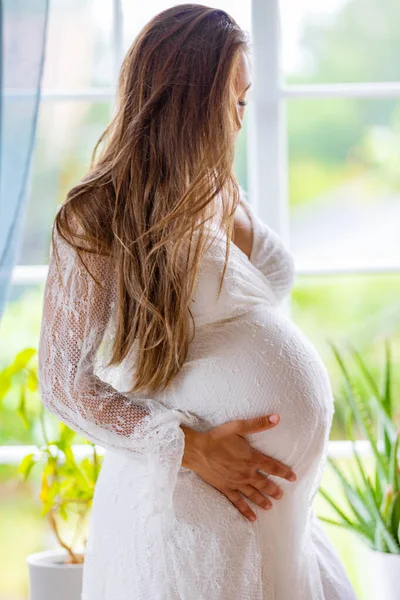 Красивая Беременная Женщина Стоит Окон Белом Платье Трогает Живот Держаться — стоковое фото