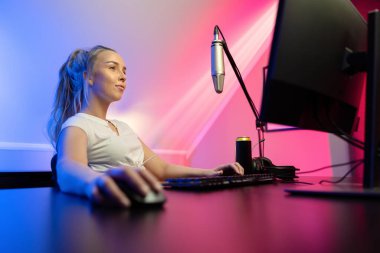Odaklanmış ve havalı görünümlü profesyonel sarışın esport oyuncusu kadın dövmeleri bilgisayarında çok oyunculu online video oyunu oynarken canlı yayında. Mavi ve pembe LED ışıklar.