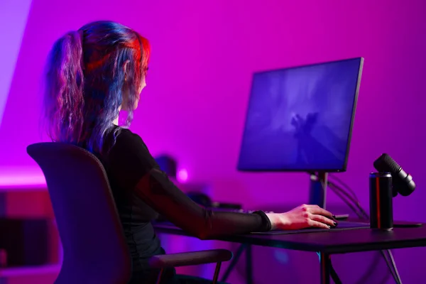 Focused professional e-sport gamer woman streaming live while playing online first-person shooter video game on her PC. Gaming room with purple and pink colored LED lights.
