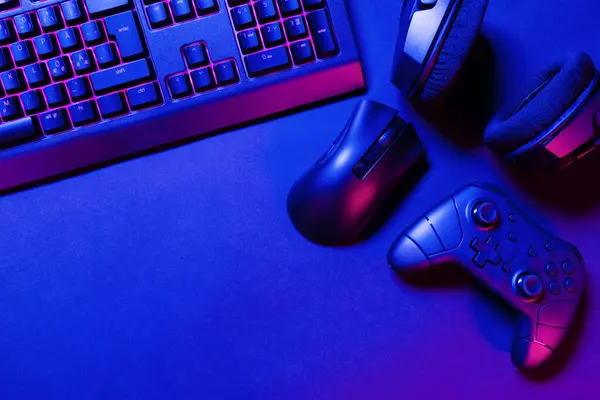 Overhead view of pink lit keyboard by game controller and mouse with headphones on black desk under blue illuminated light