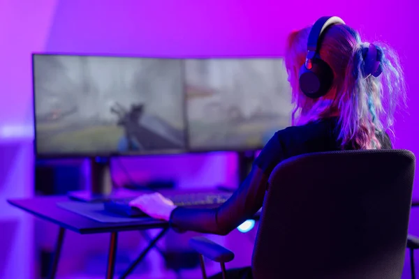 Focused professional e-sport gamer woman streaming live while playing online first-person shooter video game on her dual screen PC. Gaming room with purple and pink colored LED lights.