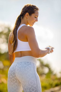 Manzaralı bir ortamda genç ve aktif bir kadın akıllı saatini kontrol ediyor. Formunu takip ediyor. Görüntü, sağlık, teknoloji ve aktif yaşam tarzını betimliyor..