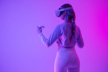 Sanal gerçeklikle ilgilenen genç bir kadın, canlı ışıklandırması olan bir odada, bir VR kulaklık ve kumanda kullanıyor. Modern teknolojiyi ve interaktif oyunları somutlaştırıyor..