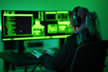 Çevresi parlayan yeşil bilgisayar ekranlarıyla çevrili siber güvenlik faaliyetlerine dalmış odaklanmış bir birey. Ayarlar gergin ve hackerların ortamını yansıtıyor..