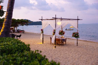 A romantic table setup under a canopy on a sandy tropical beach during sunset, accompanied by floral decorations, ocean views, and a serene ambiance perfect for a special occasion or romantic getaway. clipart