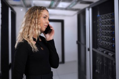 Mobil cihazla iletişim kuran profesyonel bir kadın, modern ekipmanlarla yüksek teknoloji sunucu odasında, teknolojiyi, bilişim yönetimini ve dijital altyapı bağlantısını sembolize ediyor..