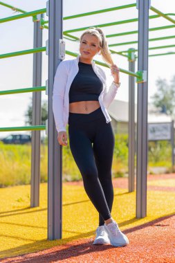 Spor kıyafeti giymiş bir kadın açık hava spor salonunda güneşli bir günün tadını çıkarıyor. Canlı bir ortamda modern fitness uygulamalarını vurgulamak.