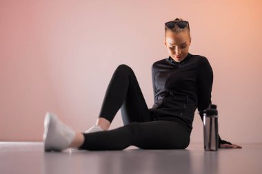 Aktivite giysisi giyen bir kadın yerde dinleniyor ve su şişesini kullanıyor. Sahne, yumuşak ışıklandırma ve rahatlama ve fitness odaklı yaşam tarzını vurgulayan sessiz bir arkaplan üzerine kurulmuştur. Kıyafeti ve duruşu sağlığını temsil ediyor.