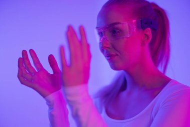 Genç bir kadın, parlak mor ve pembe ışıklı bir ortamda fütürist gözlük takarak sanal bir arayüzle etkileşime giriyor. Sahne yenilik ve teknoloji öneriyor, ileri görüşlü bir atmosferi yakalıyor. Odaklanmış ifadesi,