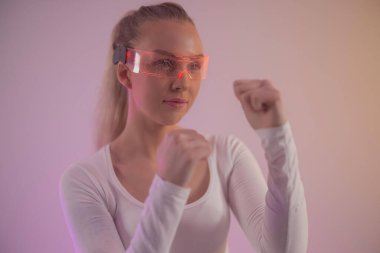 Genç bir kadın yüksek teknolojili parlak bir güneşlik takıyor ve yumruklarını boks pozunda kaldırıyor. Arka planda, fütüristik ve dinamik bir atmosfer oluşturan sıcak değişken tonlar yer alıyor. Kararlı ifadesi güç veriyor ve...