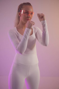 Beyaz takım elbiseli bir kadın fütürist gözlük giyerken kendine güvenerek poz verir. Canlı mor ışık atmosfere modern ve gerçeküstü bir dokunuş katıyor. Duruşu güç ve kararlılık yayıyor. Güçlenmeyi simgeliyor.