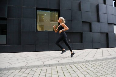 Siyah atletizm kıyafeti giyen fit bir kadın, çağdaş bir binanın önünde enerjik bir şekilde koşar. Sağlıklı ve aktif bir yaşam tarzını yansıtan görüntü, hareketi, kararlılığı ve şehir düzenini yakalar..