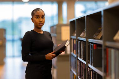 Genç bir öğrenci kütüphanedeki kitaplıkların yanında duruyor, elinde betimlenmiş bir ifadeyle bir kitap tutuyor. Sahne, sessiz bir akademik ortamda öğrenme, konsantrasyon ve entellektüel takip temasını vurguluyor.