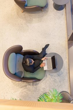 Modern ofis ortamında dizüstü bilgisayarla çalışan bir kişinin genel görünüşü. Mekan, minimalist ve sofistike bir atmosfere katkıda bulunan rahat bir koltuk, şık bir masa ve yakınlardaki bir yeşilliğe sahiptir. Uzak çalışmayı resmetmek için ideal