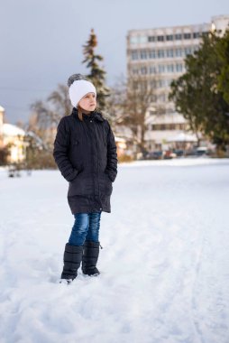 Siyah ceketli ve beyaz şapkalı genç bir kız şehrin meydanında duruyor, her yerde kar var. Kız uzun zamandır kız arkadaşını beklediği için donmuş.