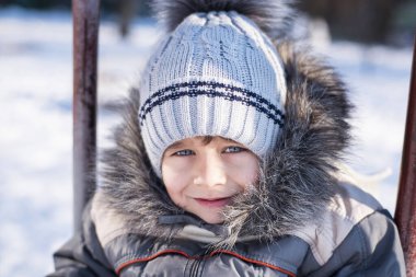 Sıcak bir şapka ve kışlık ceket giymiş güzel bir çocuğun portresi. Çocuğun etrafında her yerde kar var. Çocuk gülümser ve salıncağa biner.