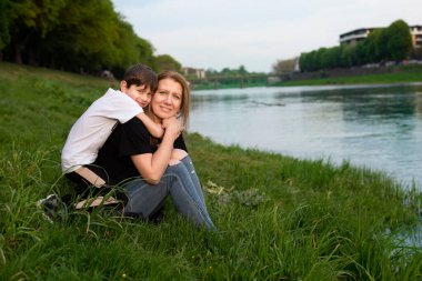 Anne ve küçük oğlu kucaklaşıp gülümsüyor. Nehir kenarındaki parkta dinlenip iletişim kuruyorlar. Anne ve oğul arkadaşlar ve birbirlerine ihtiyaçları var.