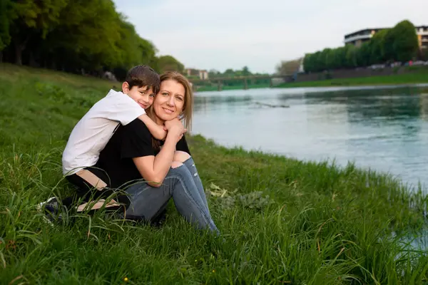 Anne ve küçük oğlu kucaklaşıp gülümsüyor. Nehir kenarındaki parkta dinlenip iletişim kuruyorlar. Anne ve oğul arkadaşlar ve birbirlerine ihtiyaçları var.