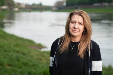 Nehrin kenarındaki parkta yürüyüp dinlenen olgun bir kadın. Temiz havada yürümek bir kadına neşe ve güç verir. Spor, yoga, doğada dua. Mutlu bir kadın gülümser.