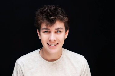 Dişlerinde metal diş telleri olan bir çocuk koyu bir arka planda açık tişörtle gülümsüyor. Bir erkek diş teli taktığı için mutlu olur ve yakında dişleri düzgün ve sağlıklı olur.