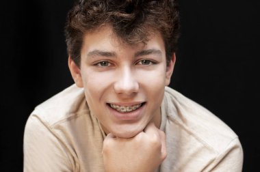 Yakışıklı genç, koyu bir arkaplan karşısında samimi bir şekilde gülümser, metal diş telleriyle dişlerinden utanmaz. Diş Konsepti