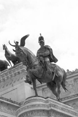 Altare della Patria 'nın bir fotoğrafı büyük beyaz mermer cephesini, yüksek sütunlarını ve Victor Emmanuel II' nin heykelini, açık mavi gökyüzüne doğru sallanan İtalyan bayrağını, hareketli Roma sokakları ve tarihi mimarla çevrili