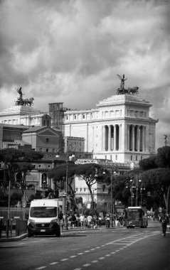 Altare della Patria 'nın bir fotoğrafı büyük beyaz mermer cephesini, yüksek sütunlarını ve Victor Emmanuel II' nin heykelini, açık mavi gökyüzüne doğru sallanan İtalyan bayrağını, hareketli Roma sokakları ve tarihi mimarla çevrili