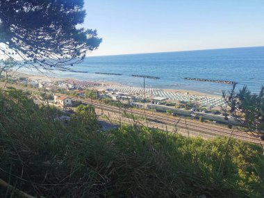 Yukarıdan çekilen bu canlı yaz manzarası Fano 'daki Fosse Sejore plajına bakıyor. Kıyı şeridi, altın kum boyunca düzgünce dizilmiş renkli şemsiyeler ve güneşliklerle dolu. Adriyatik 'in sakin, berrak suları...