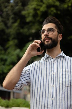 Arka planda ağaçlarla cep telefonuyla konuşan yakışıklı genç adam.