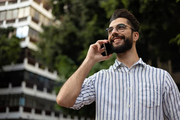 Ağaçların ve şehrin arka planında cep telefonuyla konuşan yakışıklı genç adam.