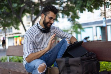 Genç sakallı bir adam açık hava bankında otururken tabletinde iletişim kuruyor.